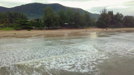 aerial shot of an island in south-east asia in which the drone is slowly moving backwards and rising up to reveal the mountains, sandy beach and a small resort in the distance