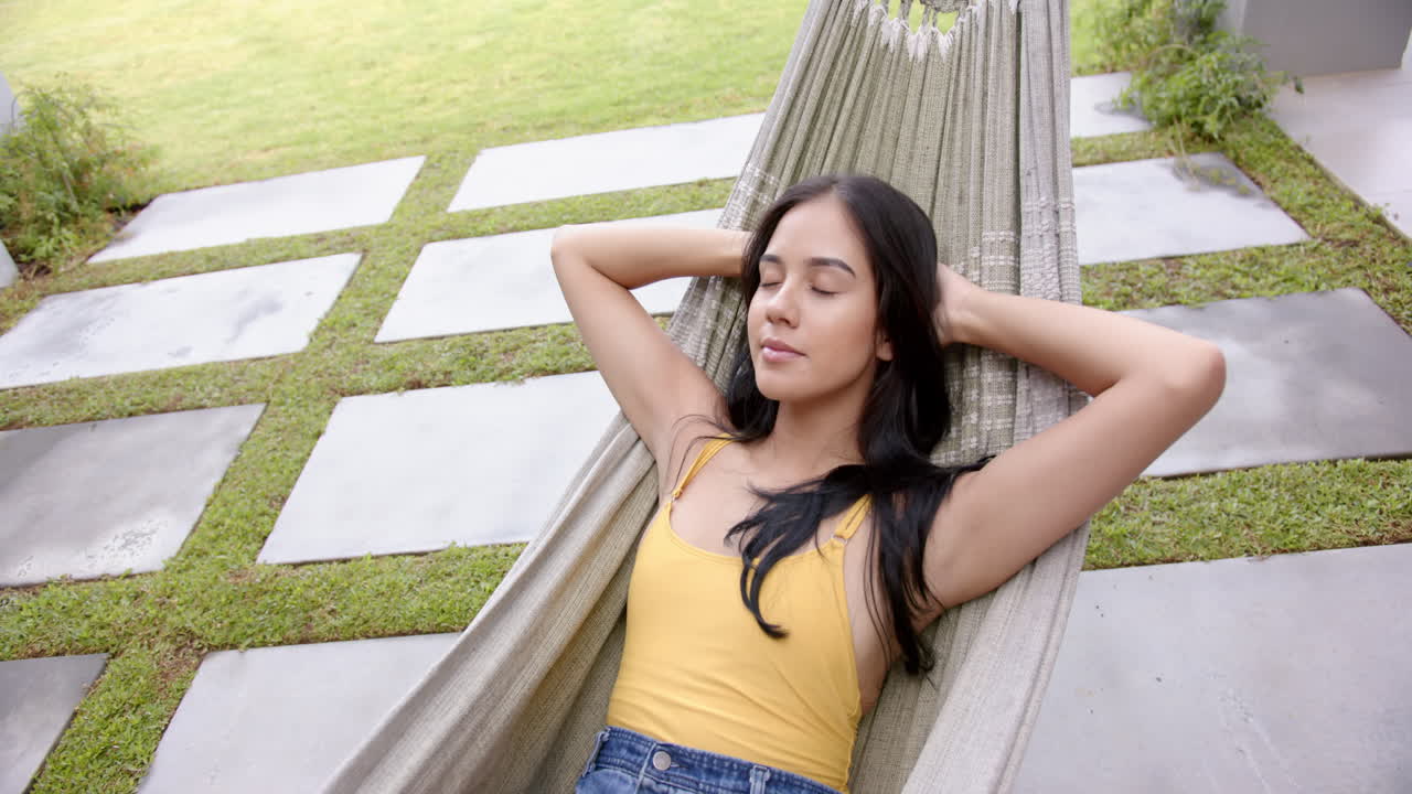 A young biracial woman relaxes in a hammock in the backyard at home, eyes closed