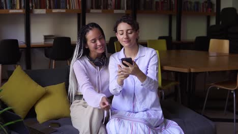 Young-Female-Students-Sitting-On-Sofa-In-Library-Surrounded-By-Books