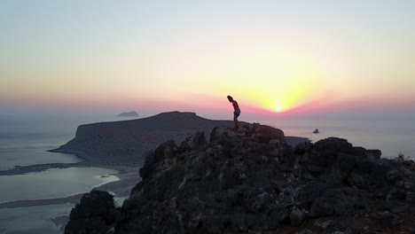 Epic-drone-footage-of-a-hiker-arriving-at-a-mountain's-peak-at-sunset-over-Balos-Beach-in-Crete,-Greece