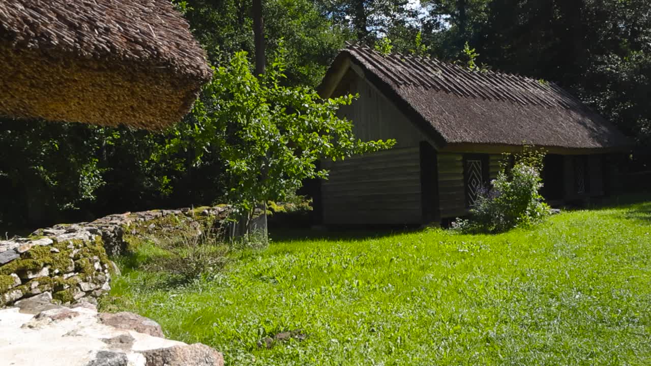Gorgeous old and traditional log farm houses or buildings with thatched straw roofing in a sunny green summer garden in the middle of a forest. Old mossy limestone fence between them, patterns on door
