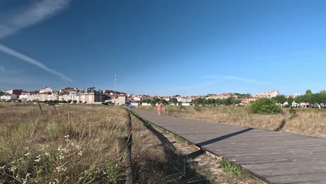 People-on-vacation-walk-relaxed-on-the-wooden-overpass-leading-to-the-city-with-the-houses-in-the-background-on-land-in-drought-on-a-bright-summer-day,-shot-blocked