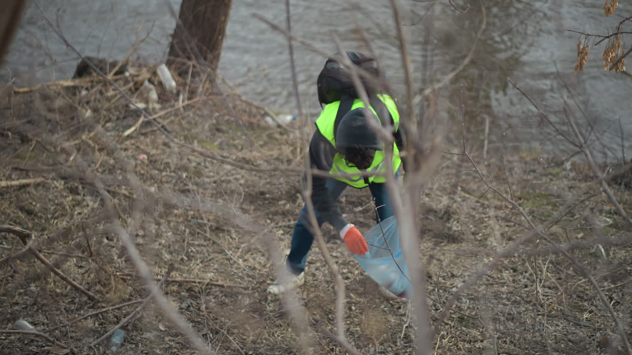 Person in reflective vest and hoodie with backpack collects trash along riverbank, placing litter into blue plastic bag during environmental cleanup effort to protect nature