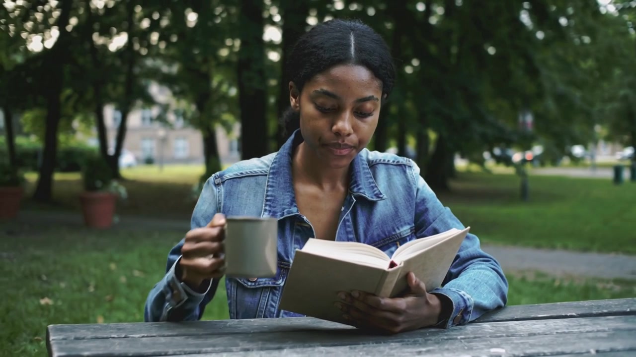 Sfogliare le pagine, donna in giacca di jeans che solleva una tazza, sorseggiando, leggendo un libro per studiare a un tavolo da picnic