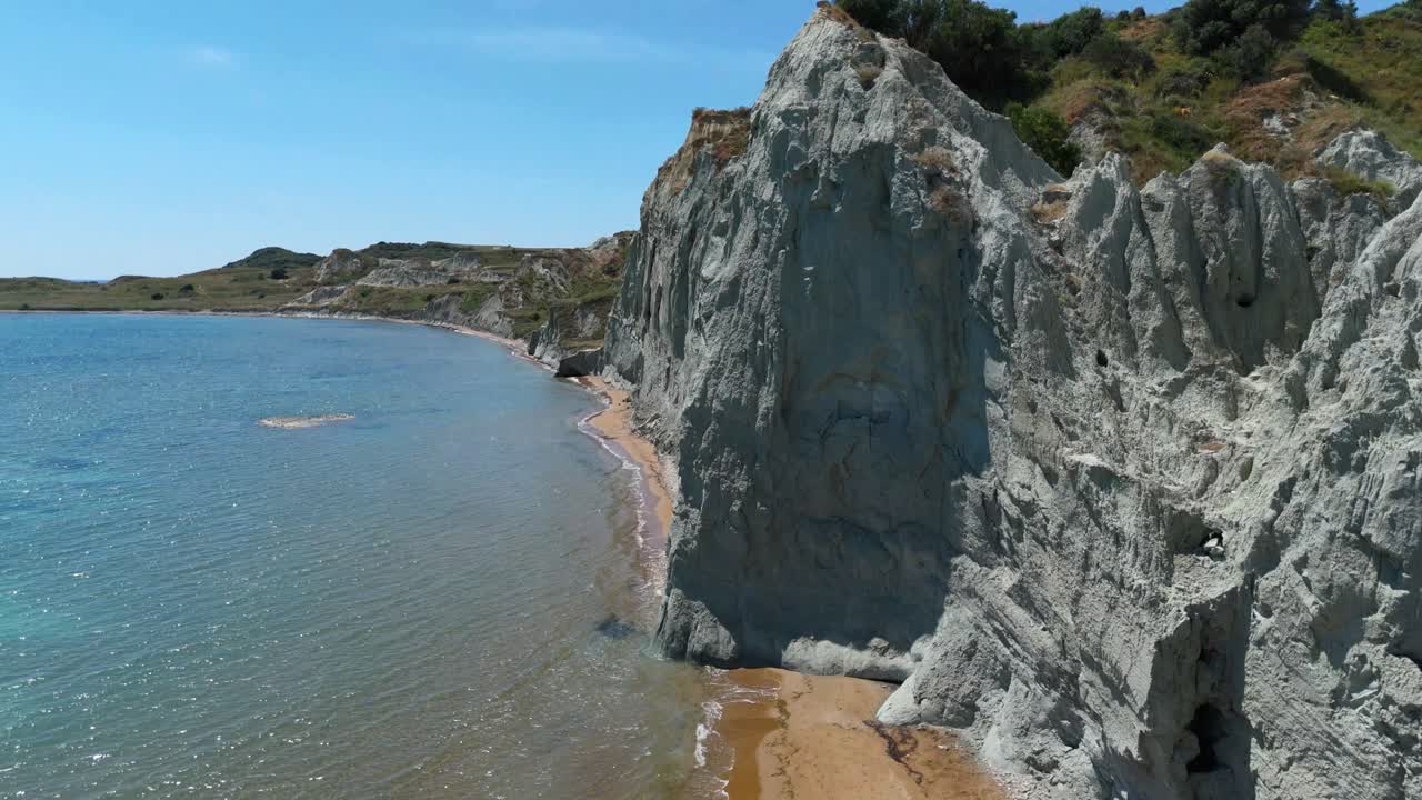 Aerial drone view of XI Beach, Kefalonia, Greece with dramatic grey clay cliffs, red sand, and crystal-clear blue waters on a sunny European travel destination.