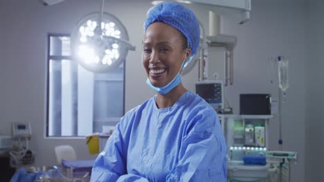 Portrait-of-mixed-race-female-surgeon-wearing-lowered-face-mask-smiling-in-operating-theatre