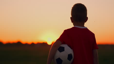 Der-Junge-Steht-Bei-Sonnenuntergang-Mit-Einem-Fußball-Auf-Dem-Feld-Im-Gras.-Er-Träumt-Von-Einer-Fußballkarriere.-Das-Konzept-Des-Erfolgs-Im-Sport.