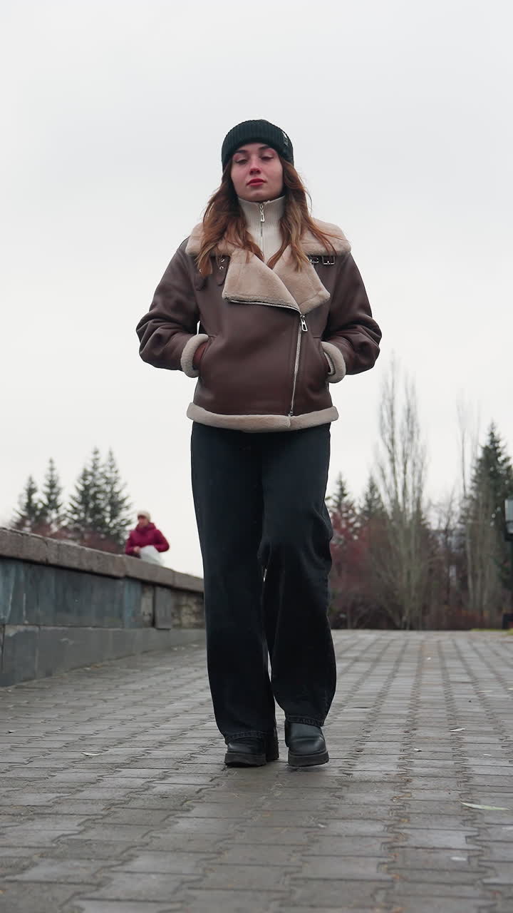 Thoughtful girl in black cap brown jacket and trousers walking slowly hands in pockets on paved pathway surrounded by pine trees and urban buildings under cloudy sky during cold peaceful autumn day