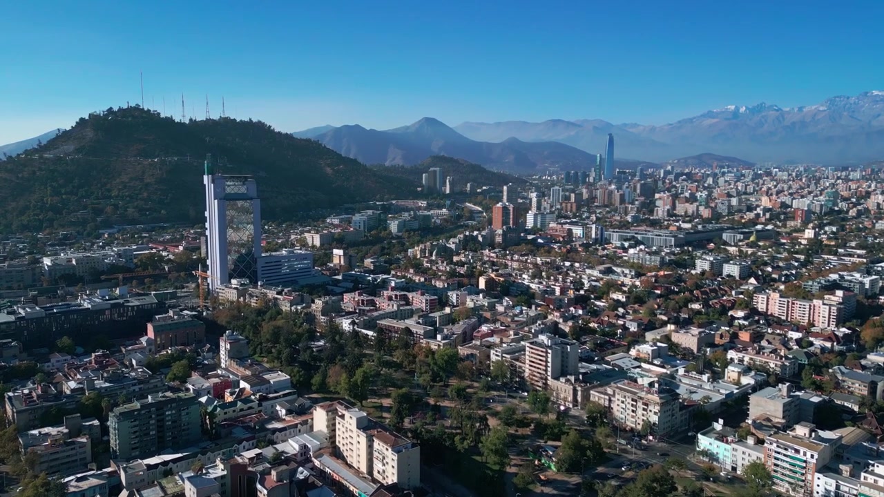 Volo sopra quartieri residenziali a bassa densità, in avvicinamento al Cerro San Cristobal, con lo skyline completo della città e le Ande innevate sullo sfondo. Santiago, Cile
