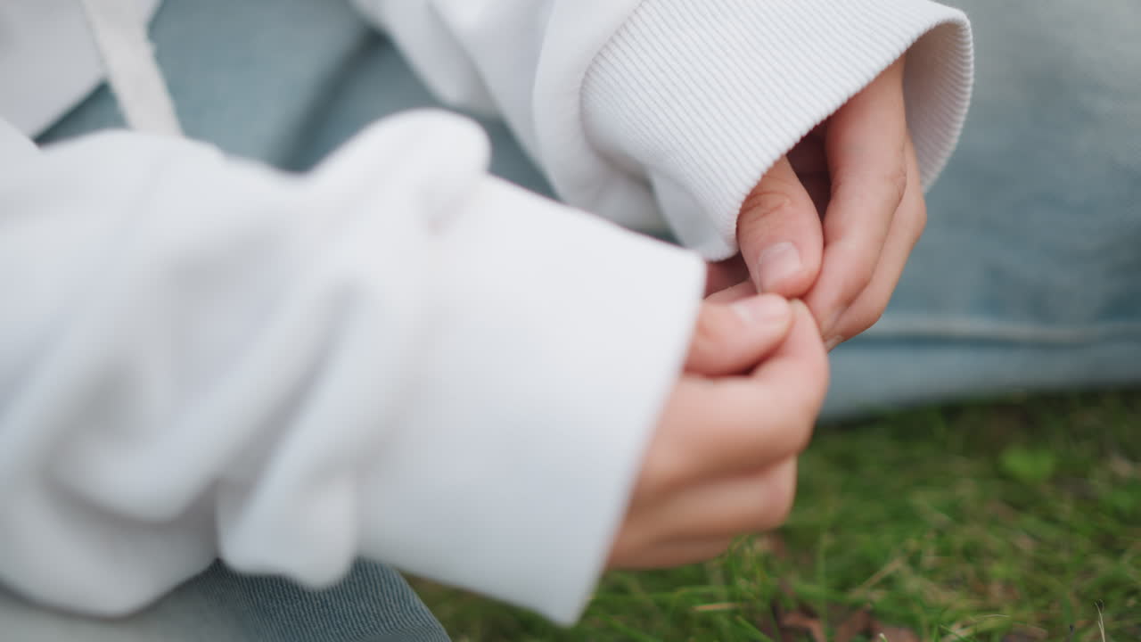 Nahaufnahme: Hände in weißem Hoodie, kleine Kinderfinger spielen mit dem Ärmel, während auf Gras sitzend. Weiches natürliches Licht, gemütliche Freizeitkleidung. Intimer, ungezwungener Moment, vermittelt schüchterne Introversion und subtile Stimmung.