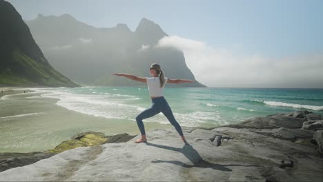 Mujer-Haciendo-Pose-De-Guerrero-De-Yoga-En-La-Playa-De-Kvalvika-En-Lofoten,-Noruega-Con-Montañas-Al-Fondo