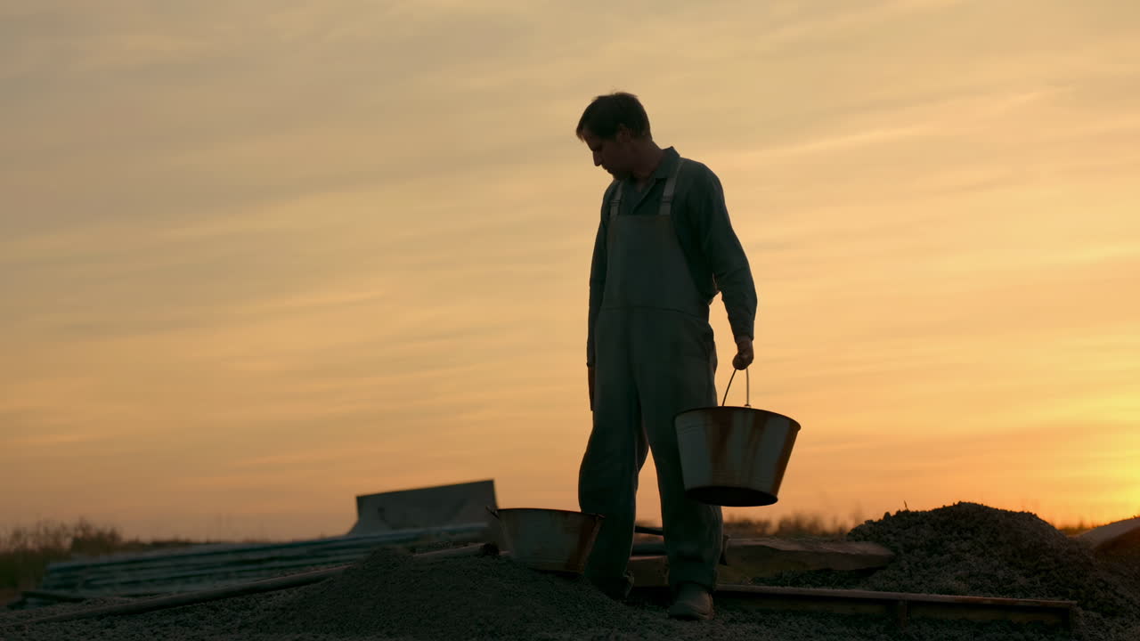 Silhouette of a Man Pouring Gravel from Bucket at Sunset