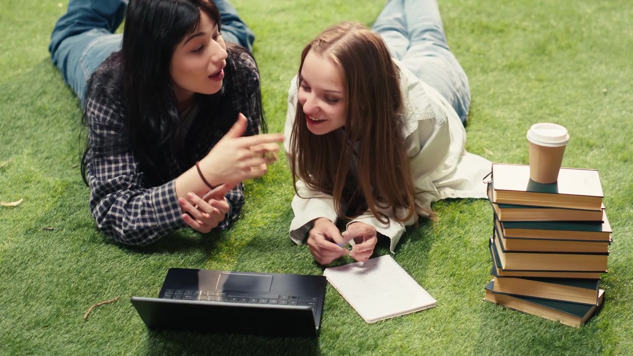 Students lay on grass beside textbook and laptop while discussing project ideas