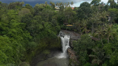 Vista-Aérea-Inclinada-De-Una-Impresionante-Cascada-En-La-Jungla-Con-Un-Hermoso-Restaurante-Al-Aire-Libre-En-La-Cima-Del-Acantilado-En-La-Selva-Tropical-De-Bali