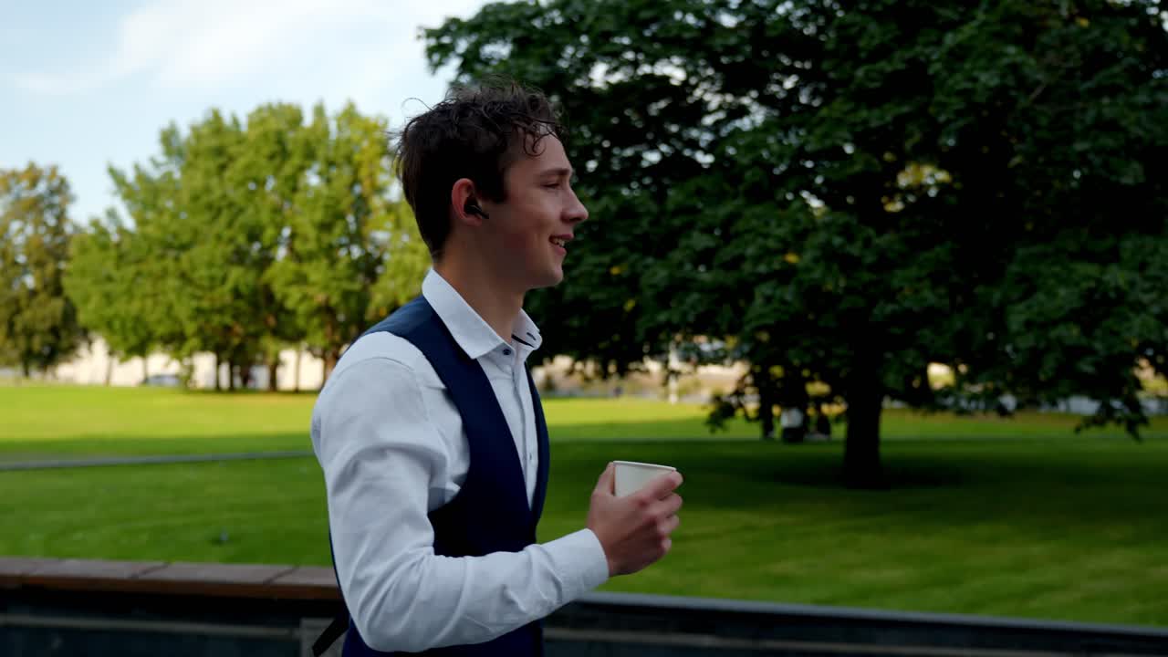 Young male in formal attire taking outdoor coffee break, enjoying a moment of relaxation while staying connected with an earbud, sipping coffee in disposable cup, slow motion follow shot