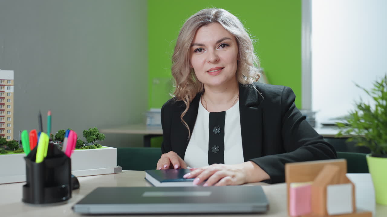 business agent closes book while smiling at desk in office with architectural model laptop and plant on table showing confident and composed demeanor in bright modern workspace