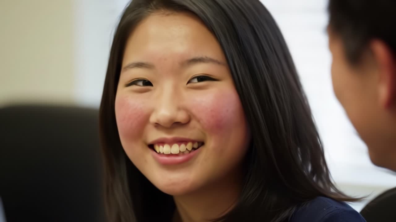 A girl with long dark hair smiles warmly while chatting with a colleague in an office setting. The relaxed atmosphere shows friendly interaction and teamwork.