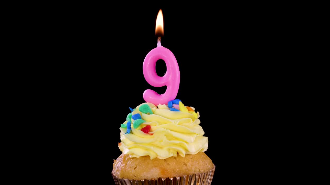 A single cupcake with yellow frosting and a lit pink number nine candle, isolated on a black background, bright studio lighting, no visible movement