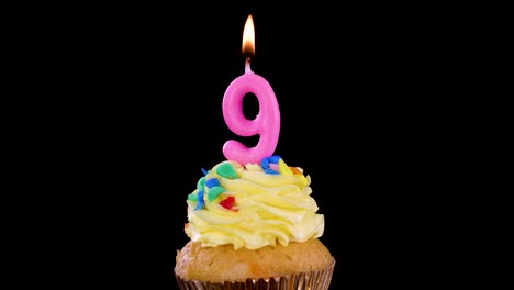 a single cupcake with yellow frosting and a lit pink number nine candle, isolated on a black background, bright studio lighting, no visible movement