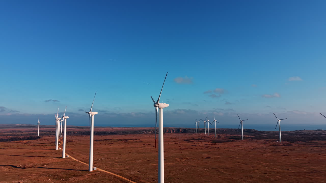 Una fila di generatori eolici che si estende all'orizzonte su terra arida e marrone sotto un cielo blu. Vista in prospettiva di turbine eoliche lungo una strada rurale.