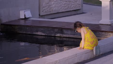 caucasian little girl sitting on the poolside watching koi fishes at resort hotel during sunset