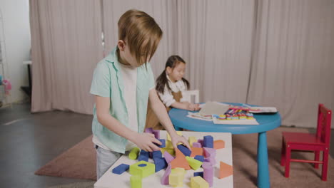 little boy in a montessori school playing with foam building blocks while little girl sitting at desk and leafing through a book