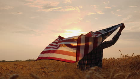 man holds usa flag behind stands in a picturesque field of wheat