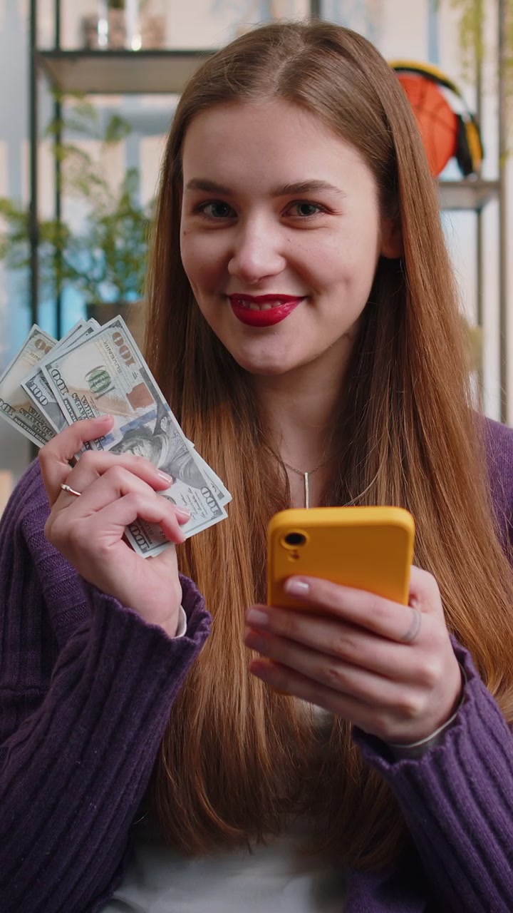 Smiling happy young woman counting money cash use smartphone income saves lottery win budget at home