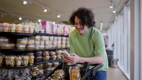 Ein-Brünetter-Typ-Lächelt-Und-Schreibt-SMS-Mit-Einem-Telefon-In-Der-Nähe-Eines-Einkaufswagens,-Während-Er-In-Einem-Supermarkt-Einkauft