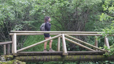 Side-shot-of-a-girl-trekking-across-a-wooden-bridge-in-a-natural-green-forest