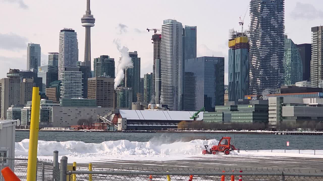 Industrial cityscape during winter with snow covered roads and distant skyline of buildings, plow pushes snow into pile as wind blows