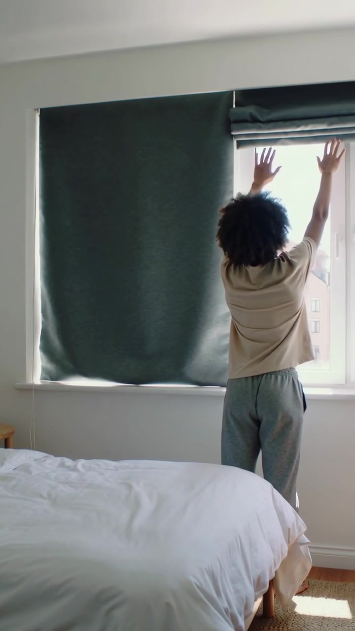 Vertical video: Pulling man in tan tee lifting roman dark shade by window, letting sun, copy space