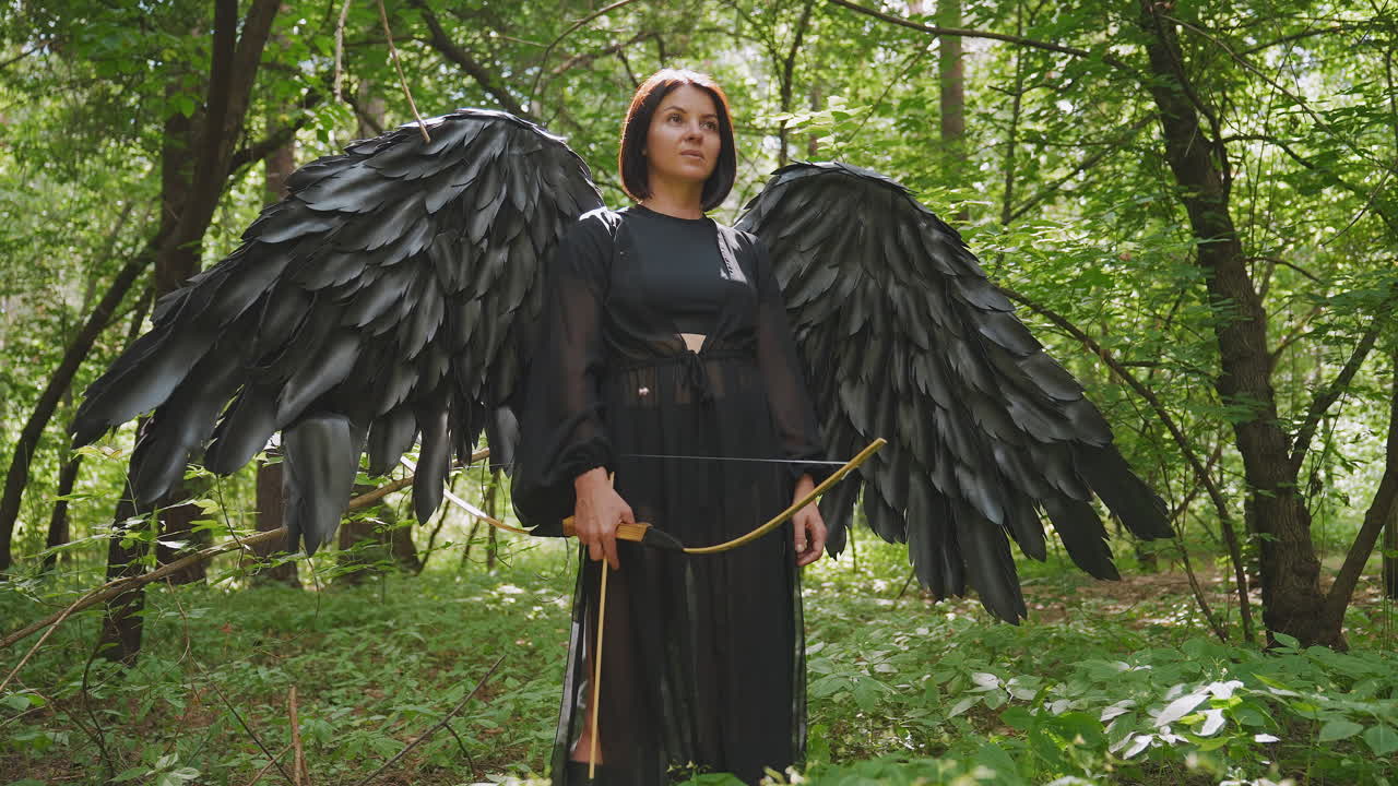 Woman with large black wings stands in green forest holding bow, sunlight filtering through leaves, serene expression reflecting power and mystery, fantasy atmosphere filled with strength