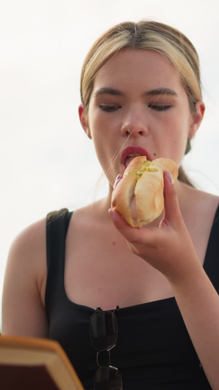 mujer con camiseta negra comiendo una hamburguesa mientras lee un libro al aire libre bajo un cielo brillante, el libro ligeramente borroso mientras se centra en la comida