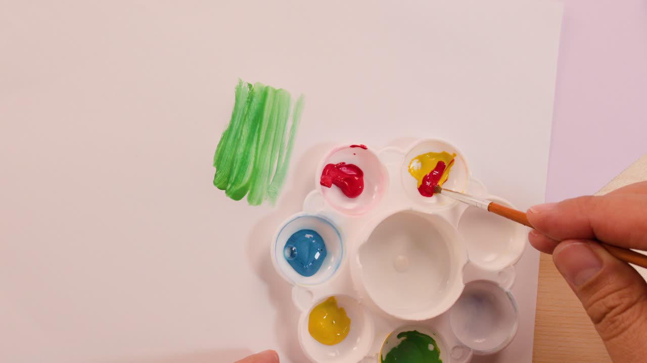 Hand mixes red and yellow paint to create orange, overhead view, bright indoor lighting