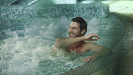 Joyful-man-resting-in-whirlpool-bath.-Smiling-man-saying-hi-at-spa-hotel.