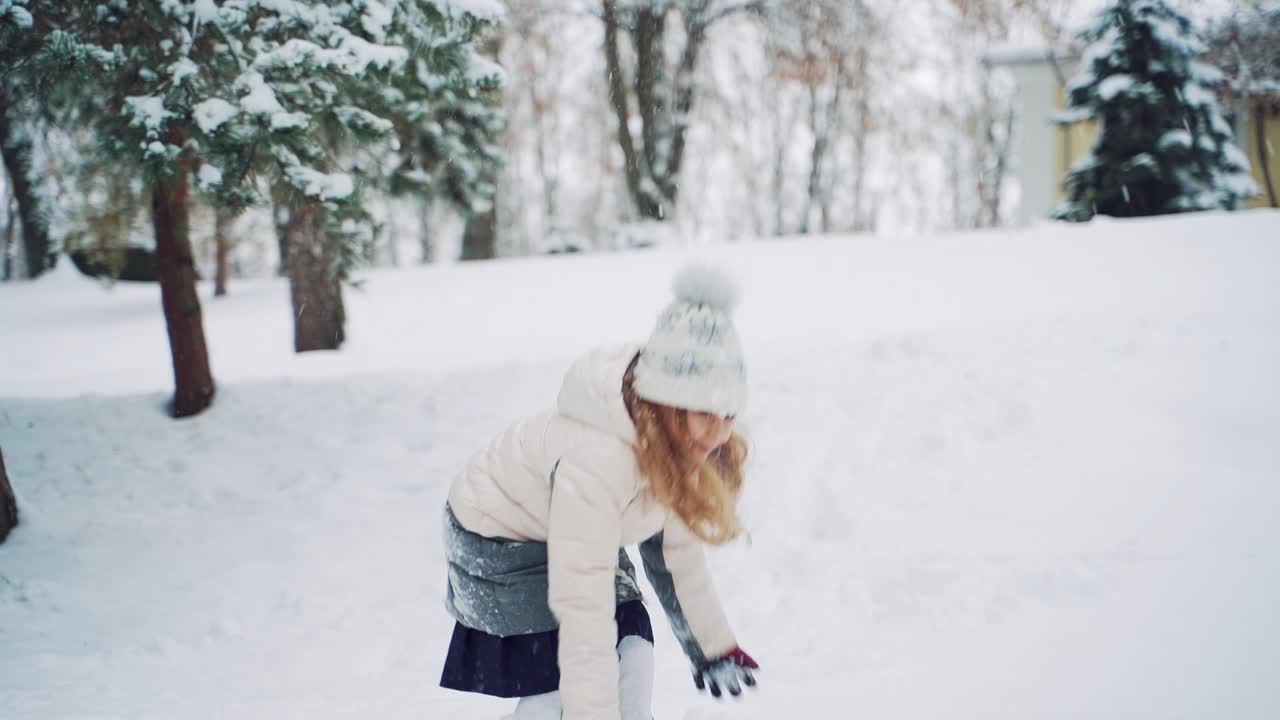 Smiling girl with long hair is throwing snowballs in the street in the yard during a snowfall in the winter. Slow motion