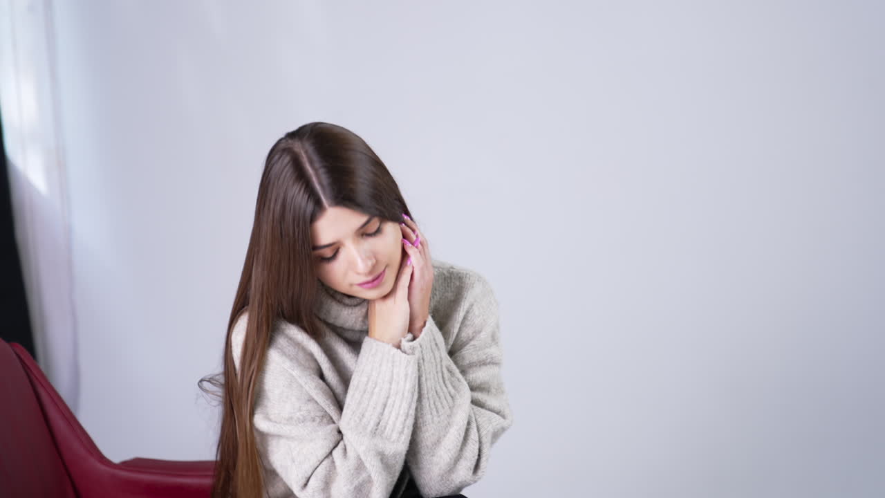Beautiful long-haired young lady sitting in armchair indoors. Attractive woman bent her head looking down. White backdrop.