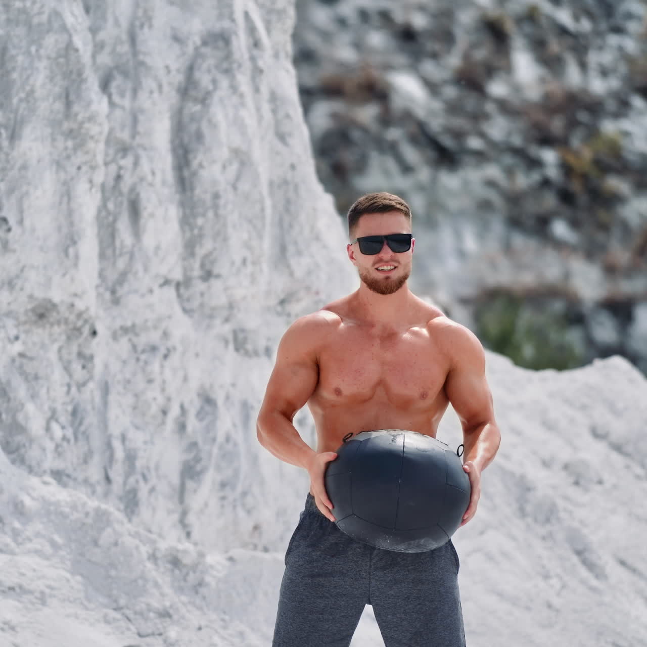 Bodybuilder training with a heavy ball in white mountains. Strong guy wearing black sunglasses doing exercises with a ball over his head outdoors in summer.