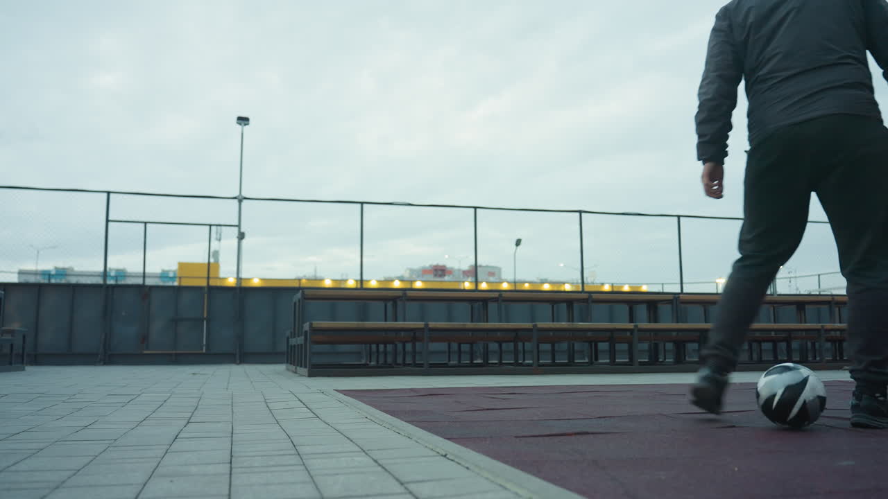 tomada amplia de una arena deportiva vacía con enfoque en bancos, valla de enlaces de cadena y alrededores urbanos, incluidos farolas y edificios lejanos con un hombre entrenando fútbol