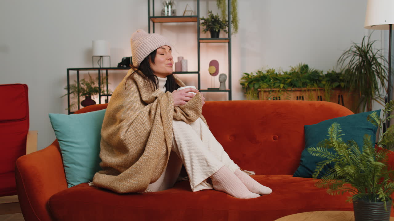병든 건강하지 않은 여성은 모자를 입고 카레드에 싸여 소파에 앉아 차가움으로 떨며 뜨거운 차를 마시고 있습니다.