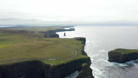 Aerial-panoramic-footage-of-sea-coast-with-high-vertical-rock-walls-against-bright-light.-Tilt-down-on-waves-crashing-on-cliffs.-Kilkee-Cliff-Walk,-Ireland