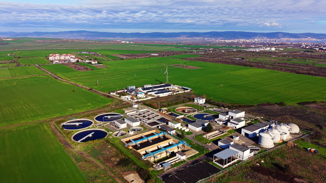 Wide drone shot of an industrial wastewater treatment facility surrounded by green agricultural fields with mountains in the distance. Bulgaria