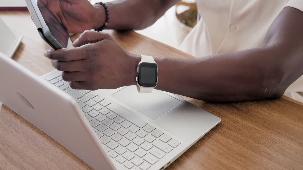Adult African American man typing on laptop at table, wearing smartwatch, pausing, checking phone