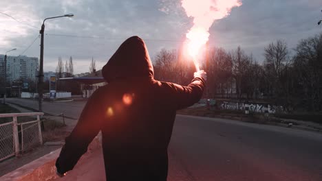 young man in hoodie and balaclava with red burning signal flare on the road under an old steel frame bridge, slow motion