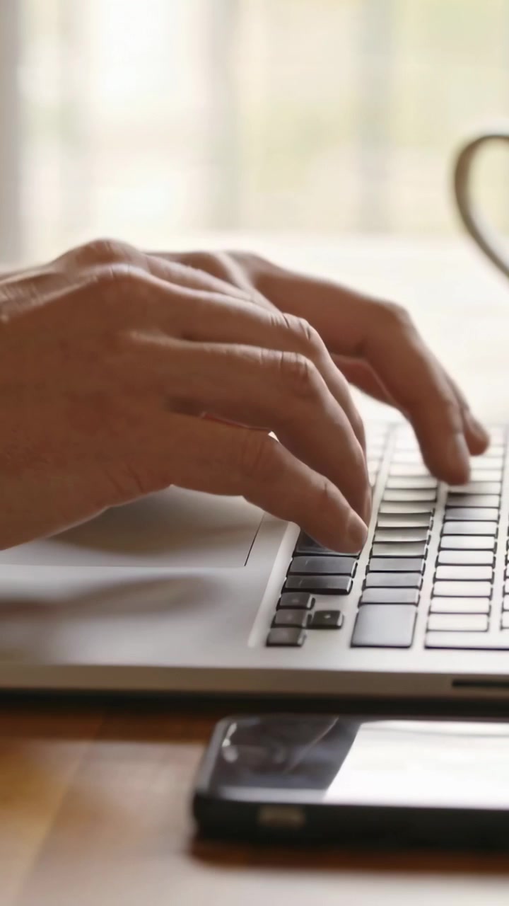 Video verticale: Dita che si librano e poi digitano, mani che lavorano al laptop sulla scrivania, con telefono e tazza