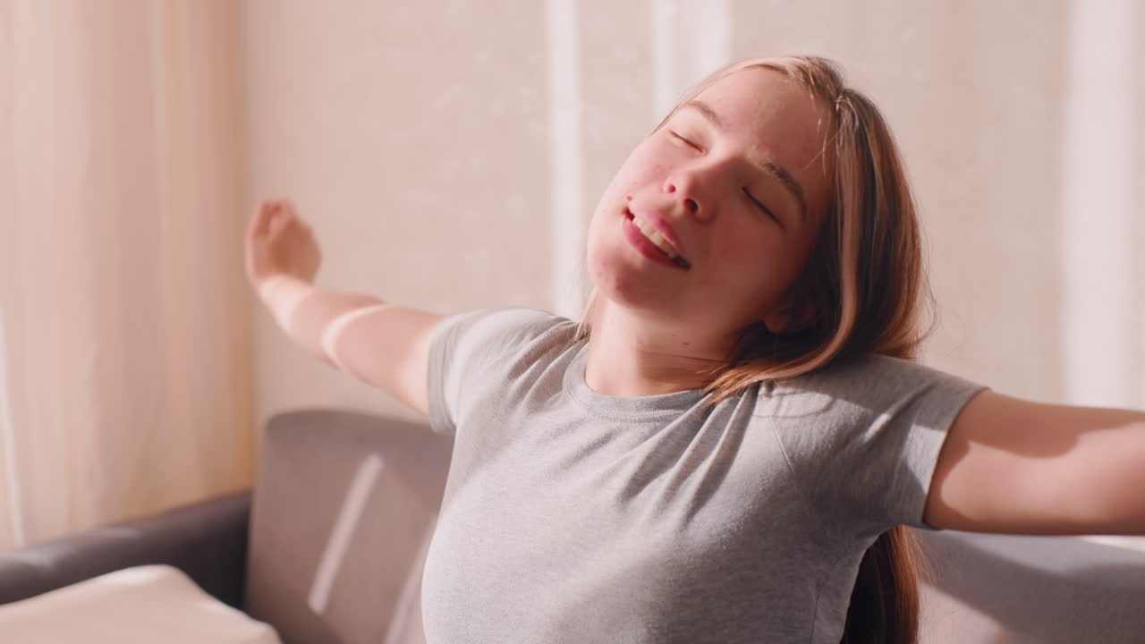 Young woman sits on couch stretching arms after waking up enjoying warm sunlight through window with relaxed smile and closed eyes creating refreshing peaceful morning atmosphere