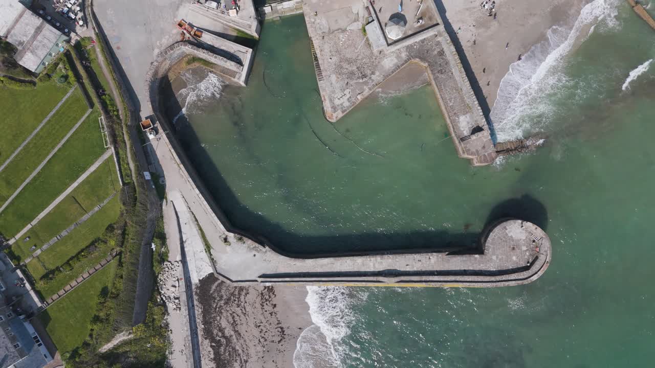 Aerial push-in over historic stone-walled Charlestown harbour with moored boats, terraced cottages and turquoise water framed by rolling Cornish hills