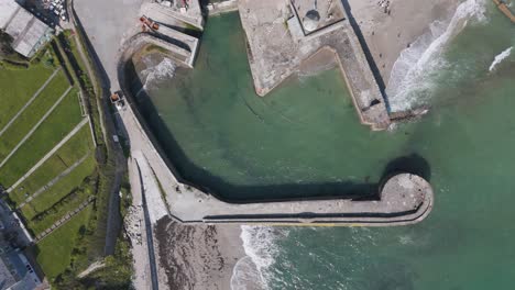 aerial push-in over historic stone-walled charlestown harbour with moored boats, terraced cottages and turquoise water framed by rolling cornish hills
