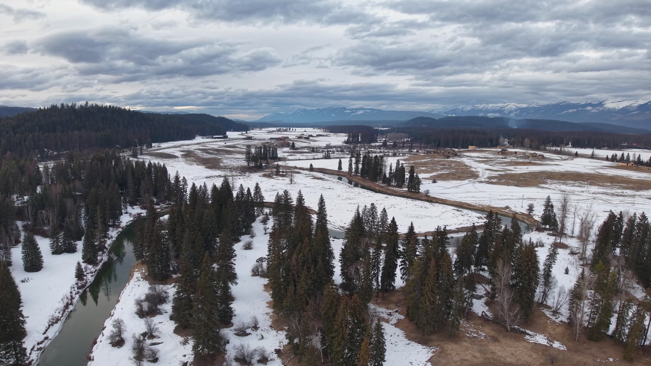 Aerial footage shows a snowy forest landscape with a winding river and scattered trees, distant mountains and a cloudy sky near Flathead River in Montana.
